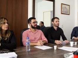vereadores sentados do mesmo lado de uma grande mesa olham para fora da foto; lemos está sorrindo; há copos de café e garrafa dágua em cima da mesa
