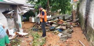 funcionário da defesa civil usando o colete laranja anota em prancheta enquanto em pé em área de lama ao lado de casas atingidas