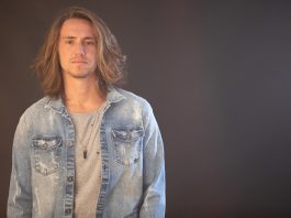 cantor victor kley posa para a câmera; ele tem cabelos loiros compridos e usa uma jaqueta jeans