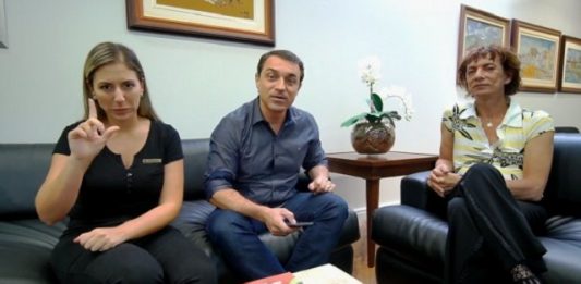 governador falando para a câmera, sentado em um sofá ao lado de uma intérprete em libras e de um homem trans