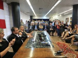 governador moisés ao lado da vice e de uma interprete em libras falando em uma mesa à frente de uma sala cheia de gente