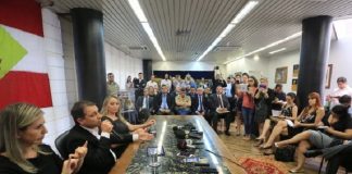 governador moisés ao lado da vice e de uma interprete em libras falando em uma mesa à frente de uma sala cheia de gente