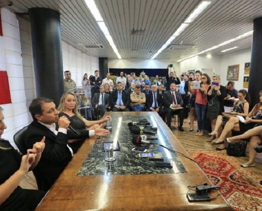 governador moisés ao lado da vice e de uma interprete em libras falando em uma mesa à frente de uma sala cheia de gente