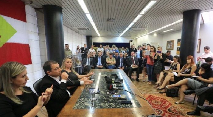 governador moisés ao lado da vice e de uma interprete em libras falando em uma mesa à frente de uma sala cheia de gente