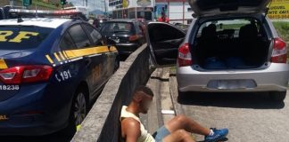 homem sentado no chão e algemado encostado no guard rail ao lado do carro furtado aberto e de viatura da prf