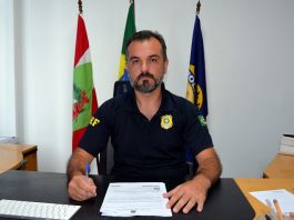 jean coelho sentado em seu gabinete olha sério para a câmera, com as bandeiras de sc, brasil e da prf atrás