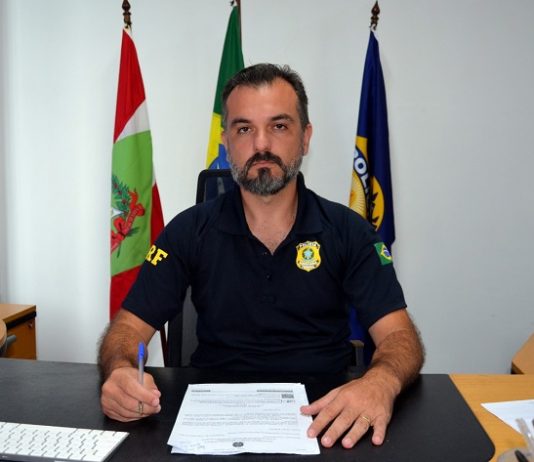 Polícia Rodoviária Federal em SC tem novo superintendente jean coelho sentado em seu gabinete olha sério para a câmera, com as bandeiras de sc, brasil e da prf atrás