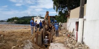 área de areia com vala aberta por escavadeira e operarios de capacete trabalhando