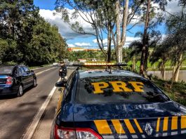 policial rodoviária parada em pé no acostamento na frente de uma viatura olhando carro passar e a estrada