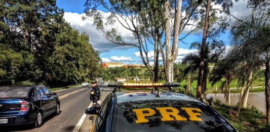 policial rodoviária parada em pé no acostamento na frente de uma viatura olhando carro passar e a estrada
