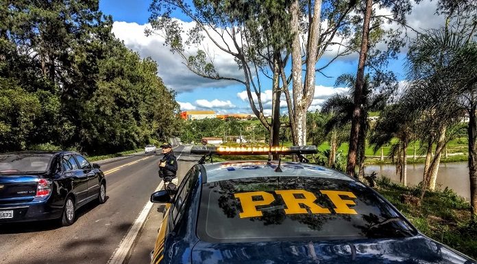 policial rodoviária parada em pé no acostamento na frente de uma viatura olhando carro passar e a estrada