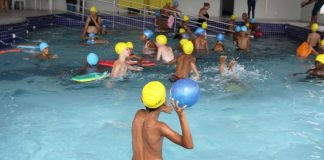 garoto visto de costas com touca de banho em piscina cheia de outras crianças; ele está prestes a jogar uma bola