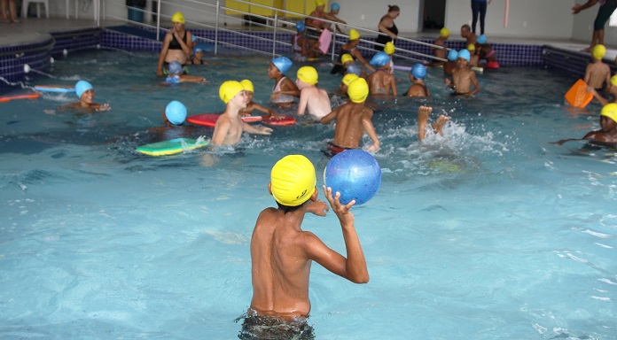garoto visto de costas com touca de banho em piscina cheia de outras crianças; ele está prestes a jogar uma bola