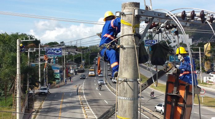 dois operários com equipamentos de proteção trabalham em ligação da rede elétrica no alto de um poste