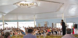 prefeita adeliana vista por detrás do palco onde há pessoas sentadas falando com a grande plateia ao fundo