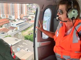 Prefeita Adeliana sobrevoa São José para verificar os estragos do temporal adeliana usando colete, óculos escuros e fone ouvido dentro do helicóptero ao lado da porta aberta olhando para fora, com a cidade em baixo; um militar ao lado