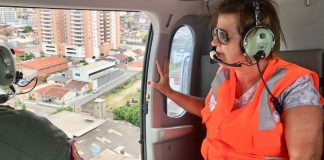 adeliana usando colete, óculos escuros e fone ouvido dentro do helicóptero ao lado da porta aberta olhando para fora, com a cidade em baixo; um militar ao lado