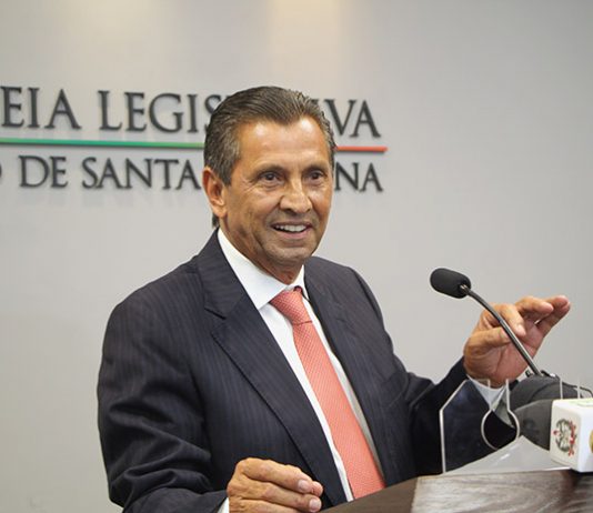 deputado fala sorridente ao microfone em bancada e atrás na parede logo da alesc