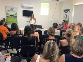 em uma sala lotada de pessoas sentadas, mulher dá palestra ao lado de um banner do ossj