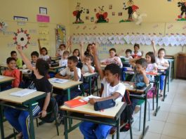 crianças uniformizadas em uma sala de aula com os braços levantados