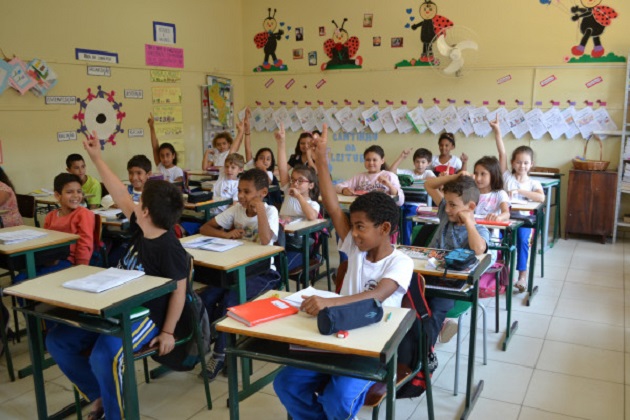 crianças uniformizadas em uma sala de aula com os braços levantados