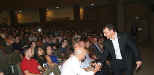 governador se inclina para cumprimentar homem sentado em poltrona do teatro, lotado de docentes