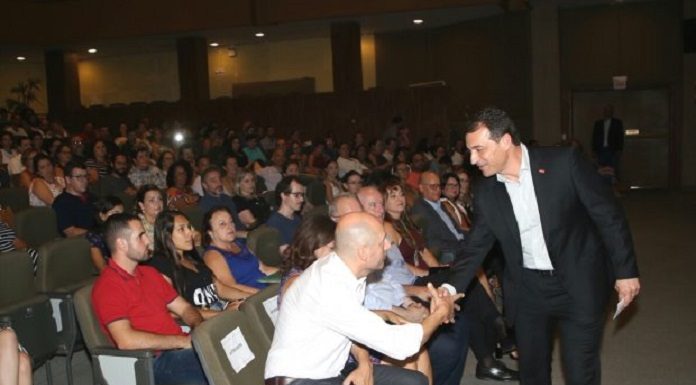 governador se inclina para cumprimentar homem sentado em poltrona do teatro, lotado de docentes