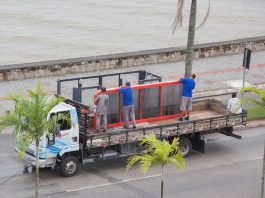 trabalhadores em cima de um caminhão colocam placa sobre um ponto de ônibus em uma avenida com coqueiros