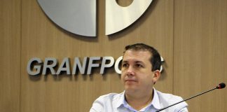 camilo martins sentado em mesa com microfone e escrito granfpolis na parede ao fundo