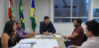 prefeito na ponta de uma mesa de mármore conversa com as demais pessoas em uma sala que tem as bandeiras do brasil, santa catarina e biguaçu