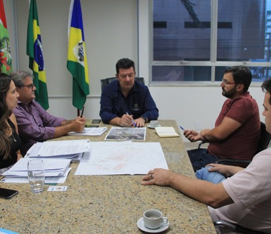 prefeito na ponta de uma mesa de mármore conversa com as demais pessoas em uma sala que tem as bandeiras do brasil, santa catarina e biguaçu
