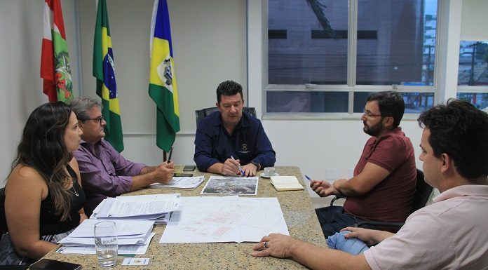 prefeito na ponta de uma mesa de mármore conversa com as demais pessoas em uma sala que tem as bandeiras do brasil, santa catarina e biguaçu