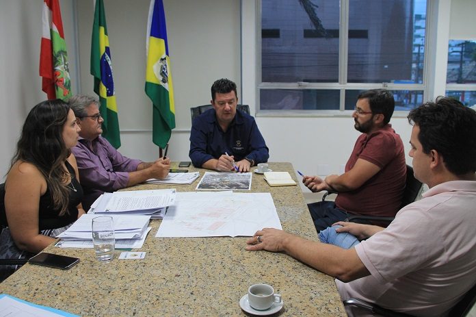 prefeito na ponta de uma mesa de mármore conversa com as demais pessoas em uma sala que tem as bandeiras do brasil, santa catarina e biguaçu