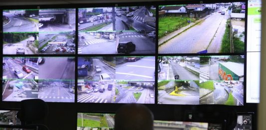 homem olhando um painel com diversas telas de cameras de ruas na central de videomonitoramento