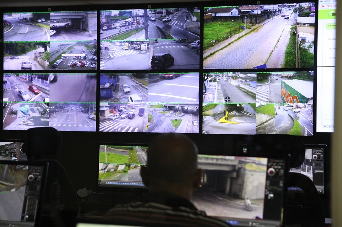 homem olhando um painel com diversas telas de cameras de ruas na central de videomonitoramento