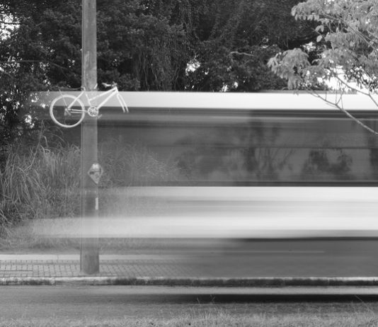 imagem em preto e branco e borrada de um ônibus passando em velocidade em frente a uma bicicleta fantasma