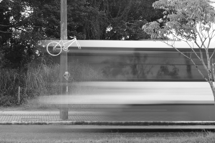 imagem em preto e branco e borrada de um ônibus passando em velocidade em frente a uma bicicleta fantasma
