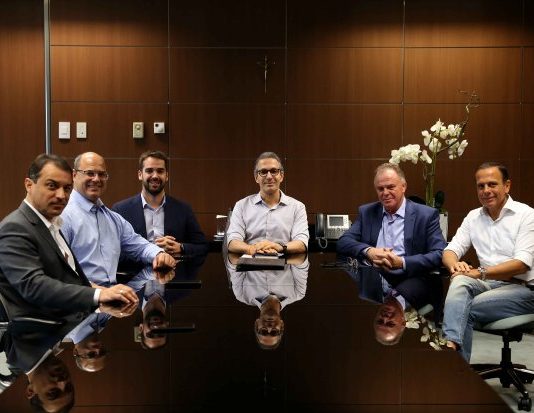 seis governadores em volta de uma mesa posa para a câmera com as bandeiras de sete estados ao fundo