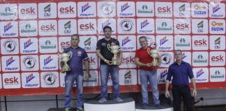 homens no podio segurando trofeus de primeiro, segundo e terceiro lugar em frente a um painel com patrocinadores