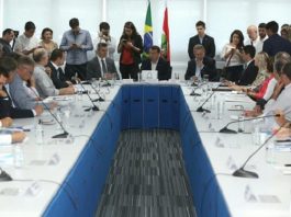 carlos moisés ao centro de uma bancada em forma de U com diversas pessoas ao redor