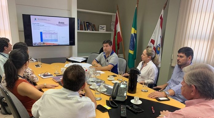 vereadores e outras pessoas reunidas em volta de uma mesa em um escritório olham para a tela de uma tv na ponta