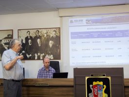 secretário fala ao microfone olhando para projeção na parede e outro homem sentado na bancada mexe no computador