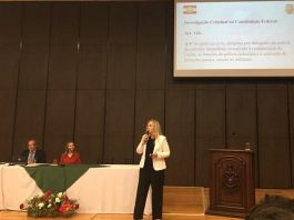 delegada em pé discursa com microfone na mão ao lado de pequena tribuna em palco com duas pessoas sentadas atrás de bancada