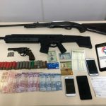 armas dispostas sobre a mesa, com munições, dinheiro, celulares e um tablet com logo da pm