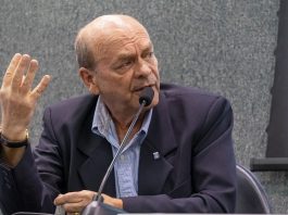 Diotallevy fala ao microfone gesticulando com a mão direita sentado em uma bancada