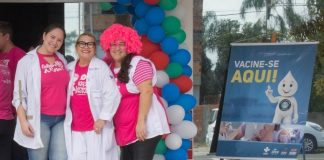 três mulheres com camisetas do outubro rosa em frete a um posto e ao lado cartaz de vacinação