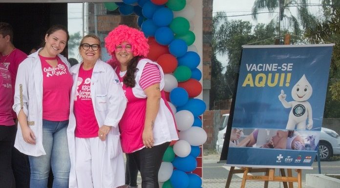 três mulheres com camisetas do outubro rosa em frete a um posto e ao lado cartaz de vacinação
