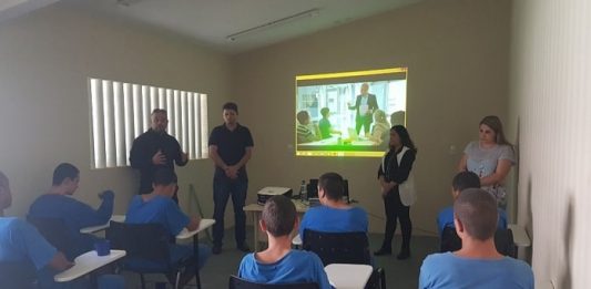 adolescentes sentados em carteiras e de costas para a câmera assistem adultos em pé falando com um projeção de slide na parede