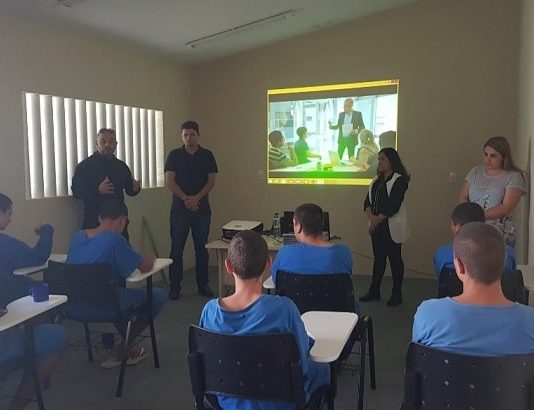 adolescentes sentados em carteiras e de costas para a câmera assistem adultos em pé falando com um projeção de slide na parede