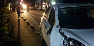 nissa march branco com parte da frente batida parado no acostamento e policias e corpo no chão ao fundo em foto noturna